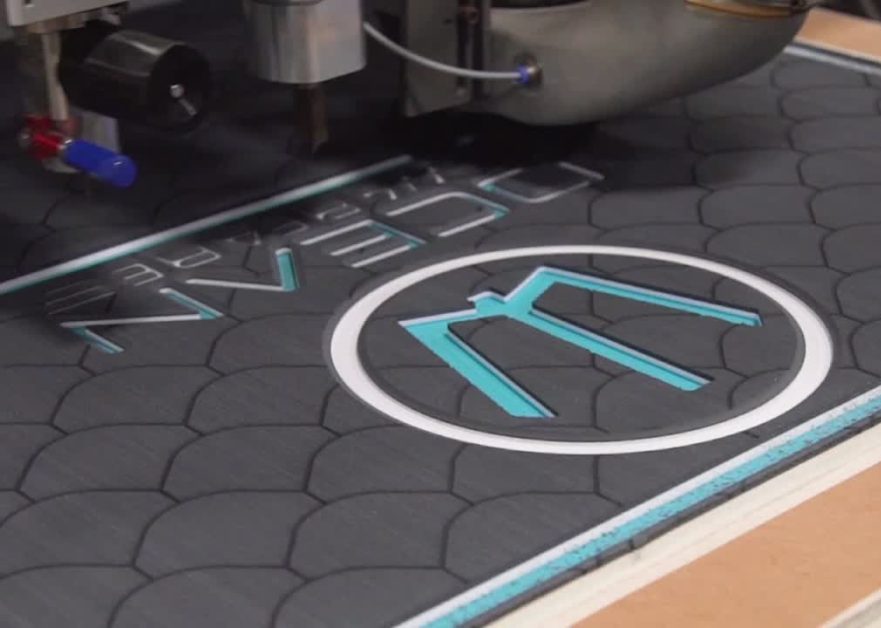 CNC machine cutting custom SeaDek marine flooring with logo and fish scale pattern in dark gray and teal